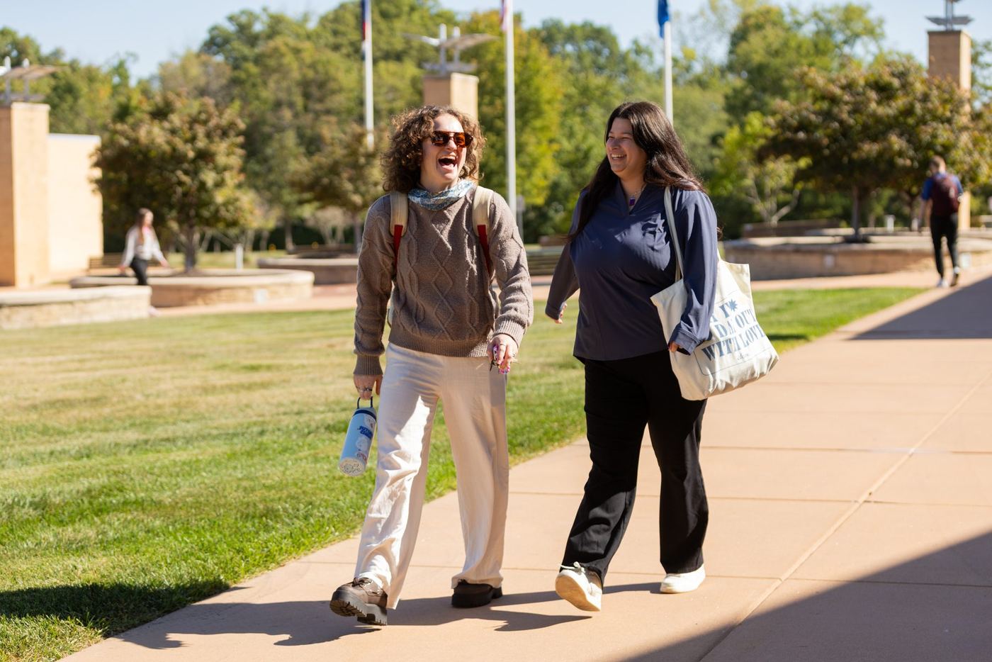 Two smiling women walk on a paved path next to grass, with campus buildings and trees in background.