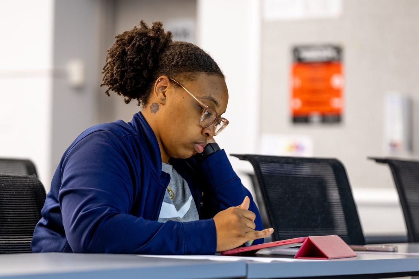 Student in blue jacket and glasses focuses on a pink tablet at a gray desk in a modern classroom.